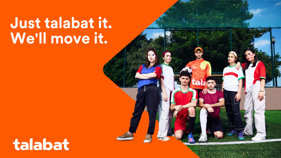 Key visual — seven young people in football shirts pose on a pitch in front of a fence, beside an orange panel with the line “Just talabat it. We’ll move it.” and the talabat logo